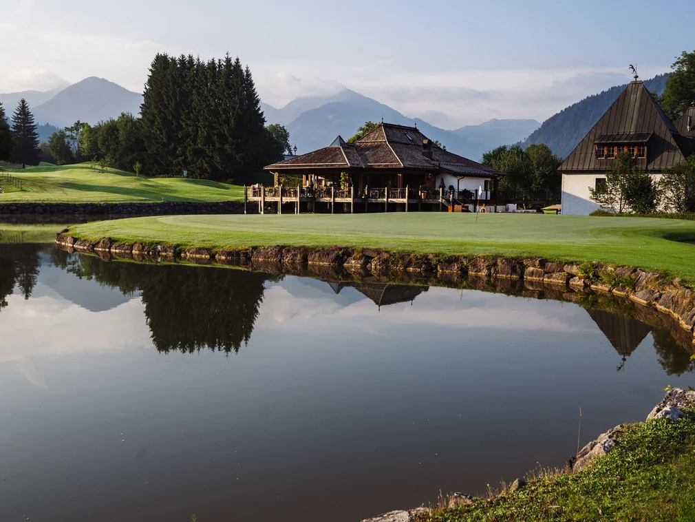 Golf course with pond, trees, clubhouse, and mountains in the background