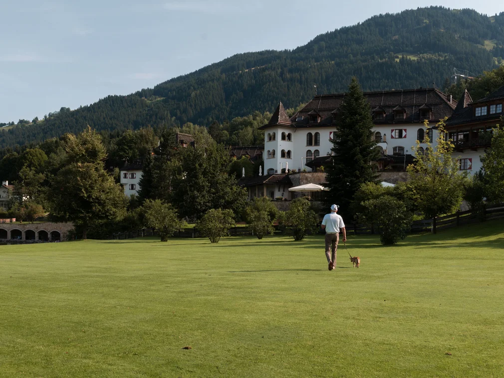 Man walking dog on green lawn in front of castle with forested mountain background