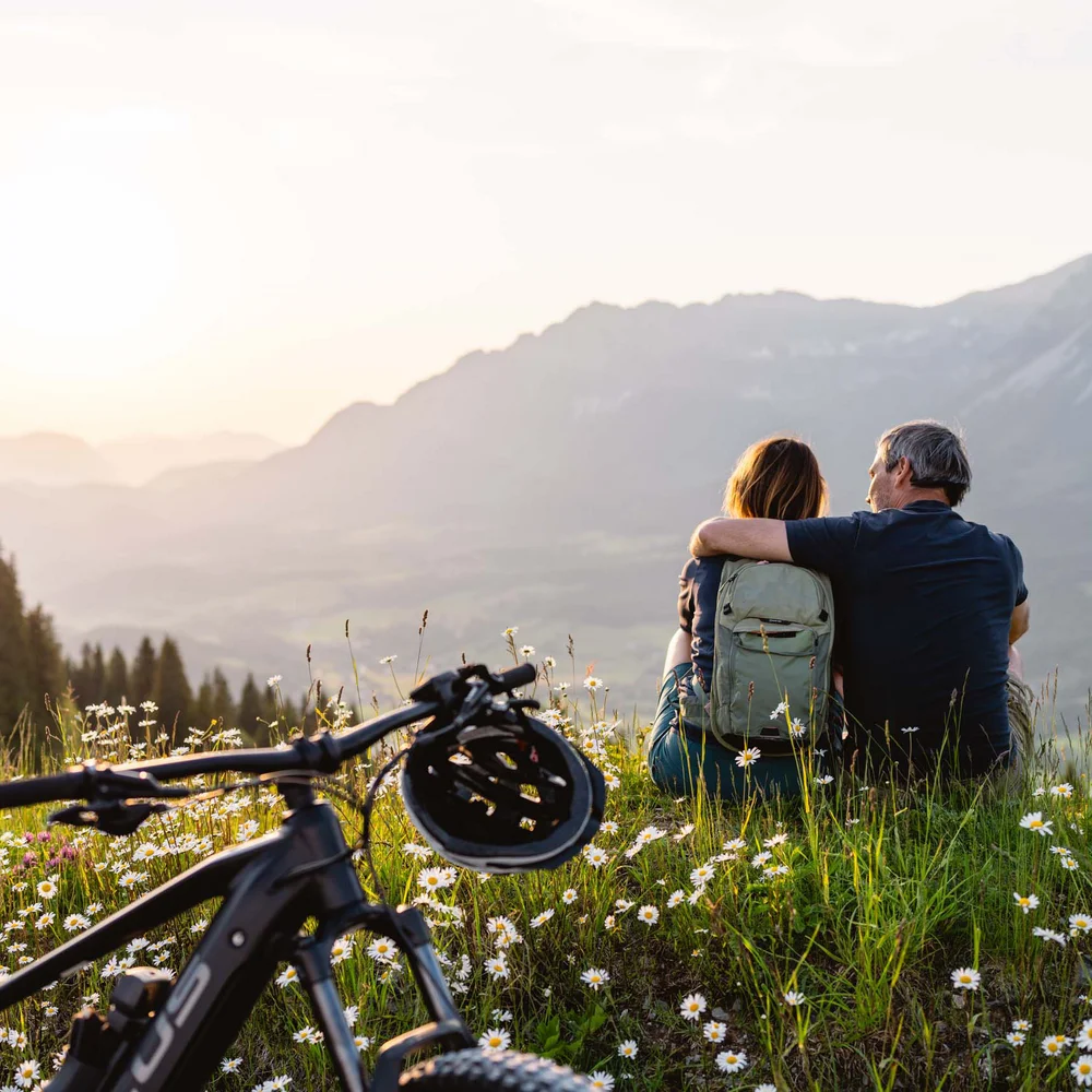 Blog Paar sitzt auf einer Blumenwiese, blickt auf Berge bei Sonnenuntergang