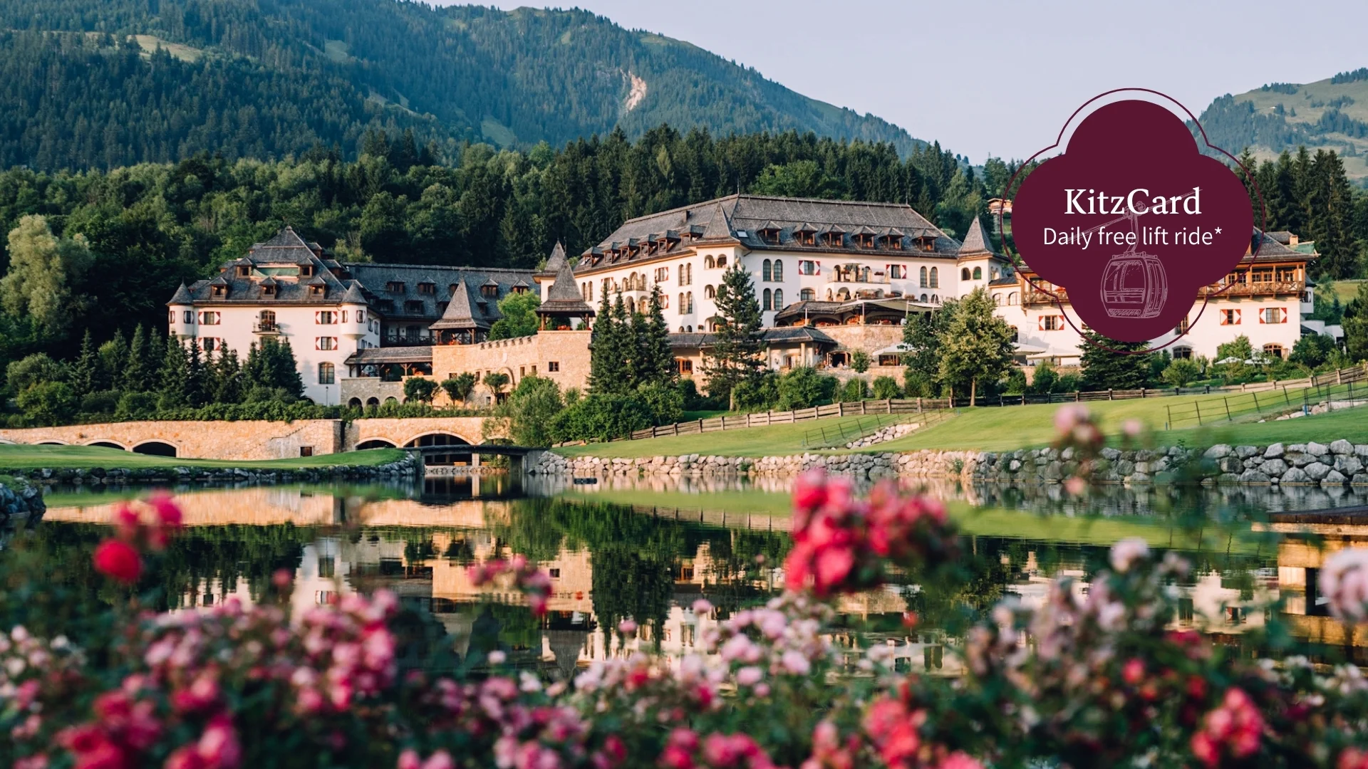 Schlosshotel Kitzbühel – a premium experience Hotel complex by a lake with mountains, bridge and blooming flowers in the foreground