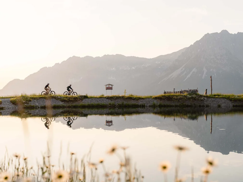 Two cyclists riding by a lake with mountains in the background at sunset