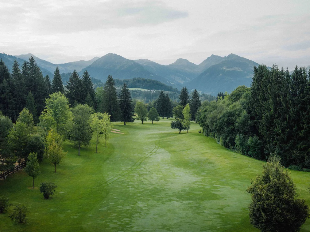 Green golf course with trees and mountains in background under cloudy sky