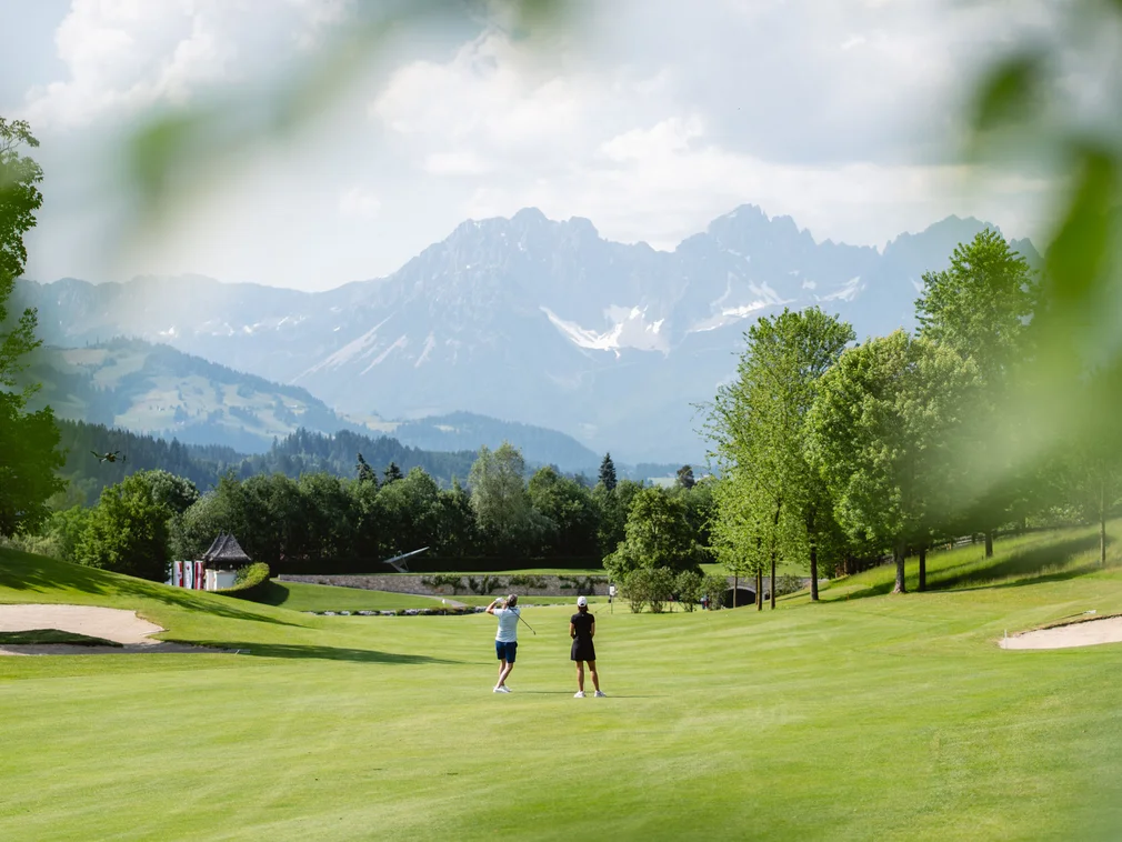 Two golfers on green golf course with mountains in background on sunny day