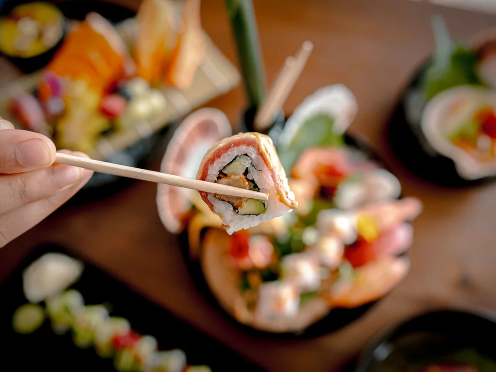 Hand holding sushi with chopsticks above plate with assorted sushi