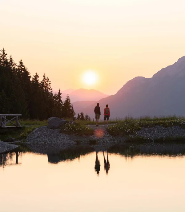 Flitterwochen im Hotel in Österreich | Schlosshotel Kitzbühel Zwei Personen betrachten den Sonnenuntergang an einem Bergsee mit Wald und Brücke