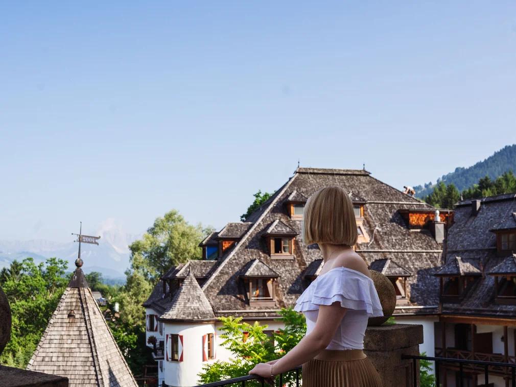 Woman on balcony looking at traditional house with mountain backdrop under clear sky