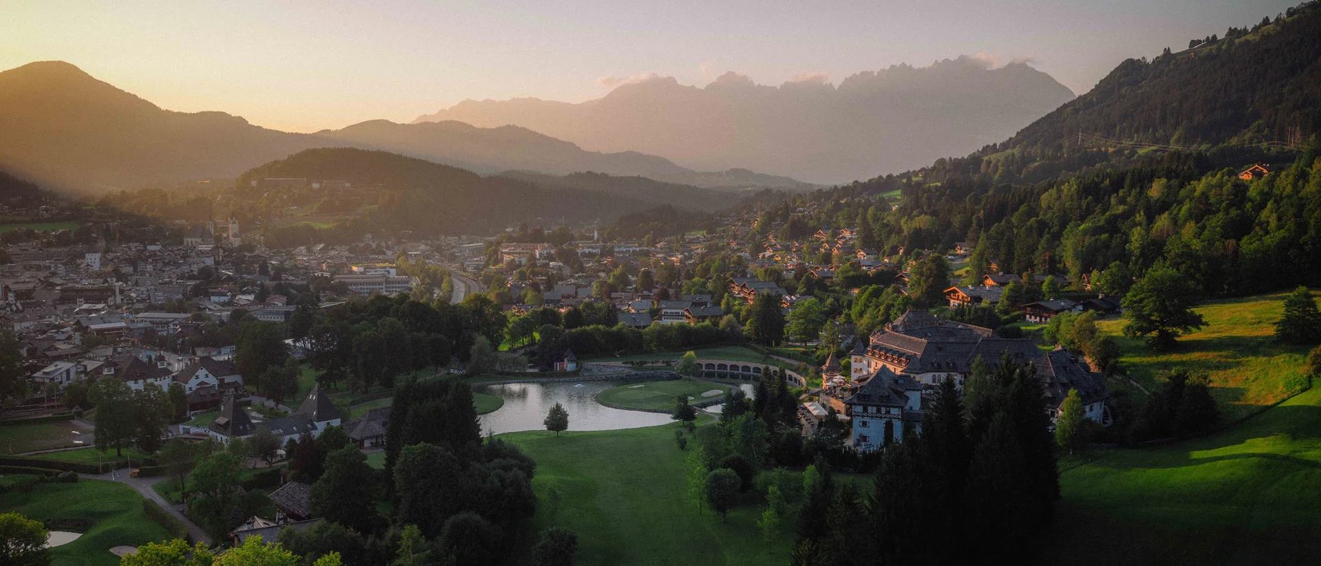 Schlosshotel Kitzbühel von oben Blick auf das Schlosshotel Kitzbühel, den Golfplatz des Golfclubs Kitzbühel und den Ort Kitzbühel aus der Vogelperspektive. Am Horizont senkt sich die Sonne hinter dem Gebirge Wilder Kaiser