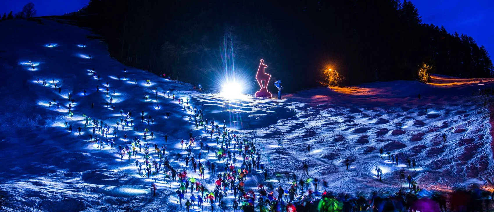 Nachtlauf mit vielen Läufern auf schneebedecktem Hang und Leuchtfigur am Waldrand