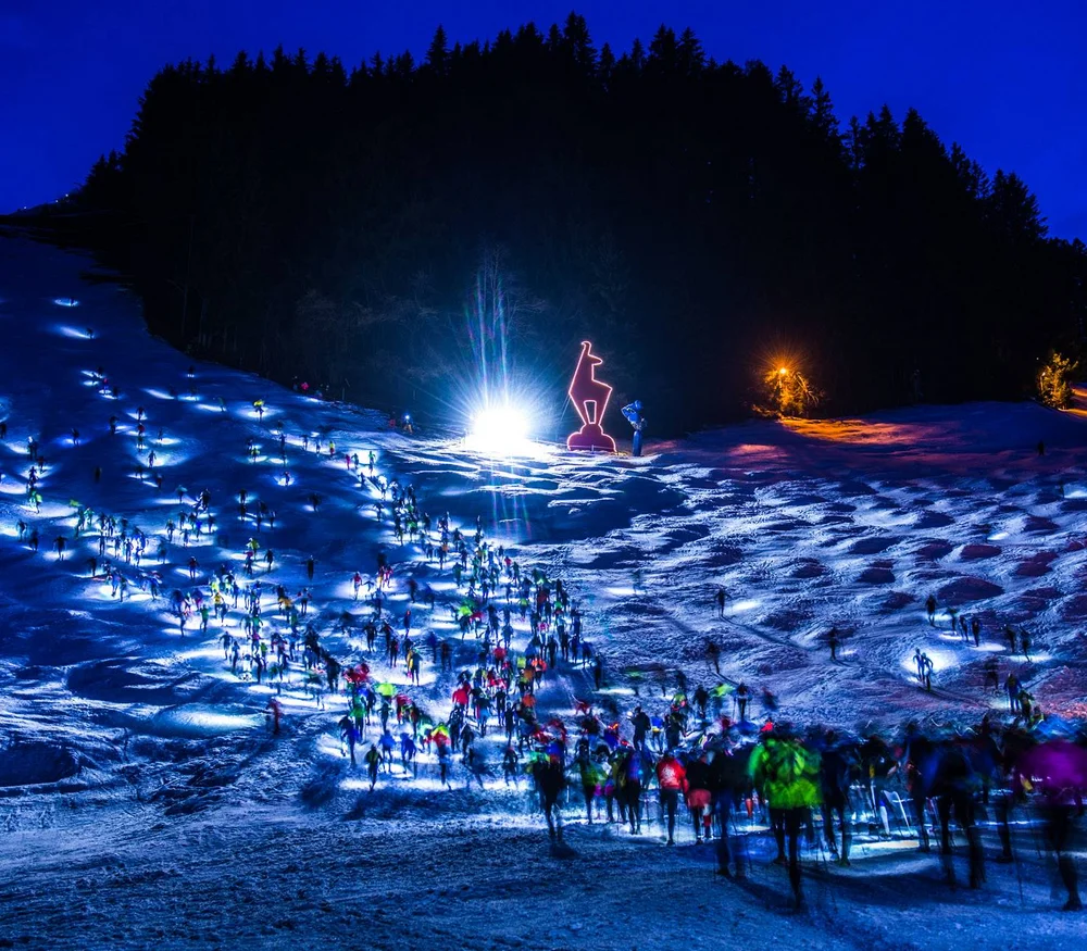 Nachtlauf mit vielen Läufern auf schneebedecktem Hang und Leuchtfigur am Waldrand