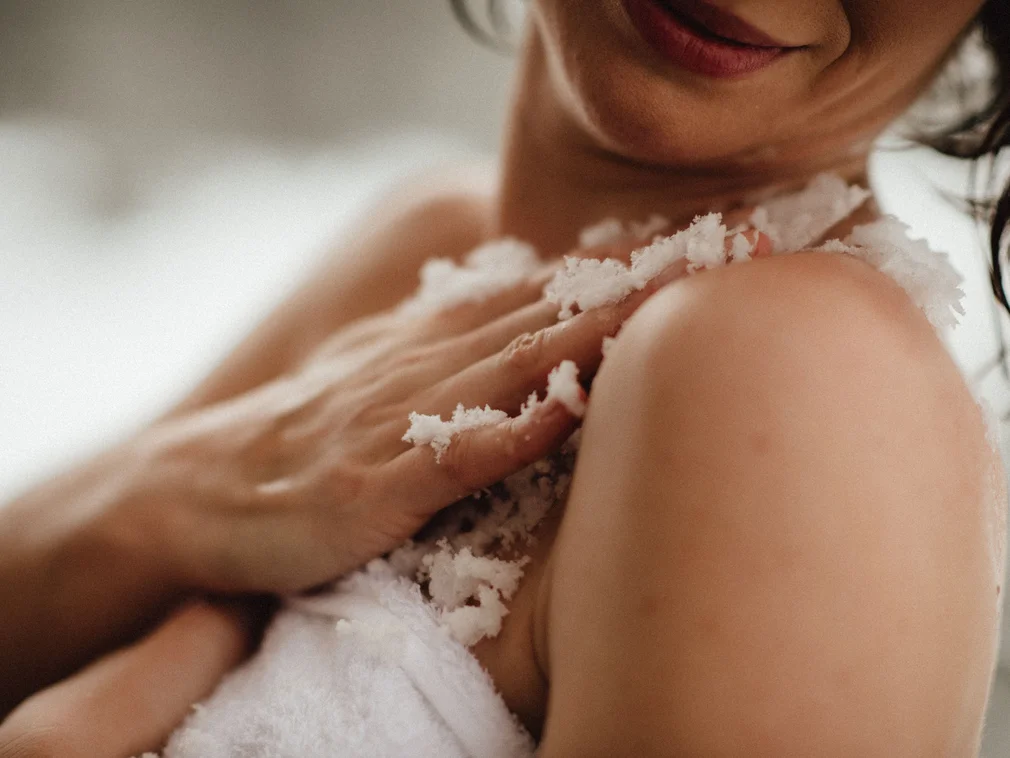 Woman applying body scrub on shoulder in bathroom