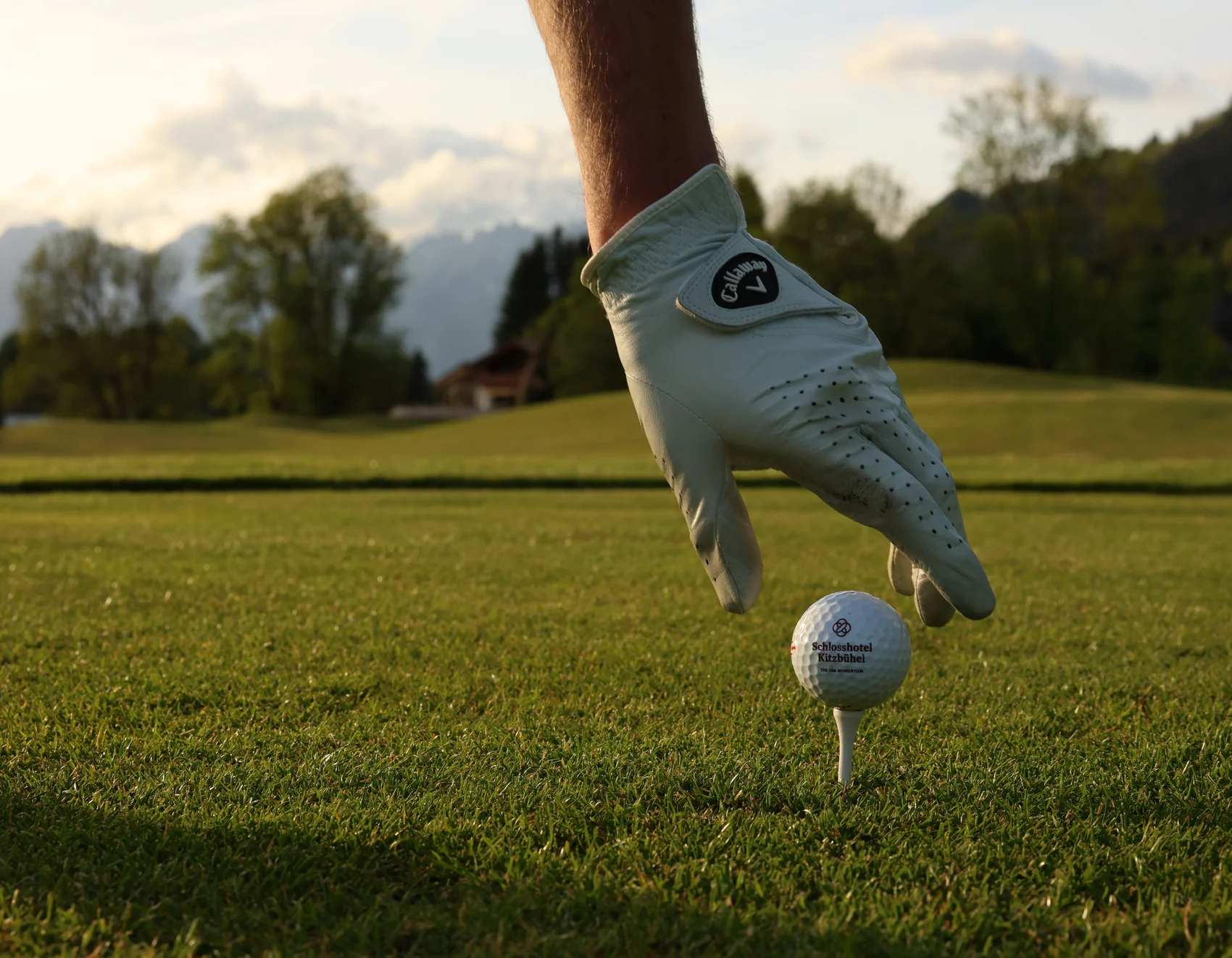 Golfspieler mit weißem Handschuh platziert Golfball auf Tee, Alpen im Hintergrund, Sonnenuntergang, Schlosshotel Kitzbühel.