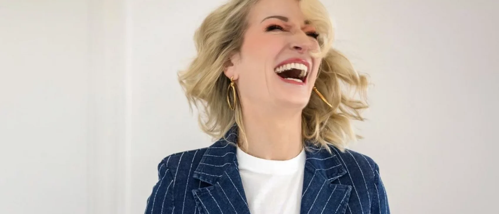 Frau mit blonden Haaren lacht laut in einem gestreiften Blazer