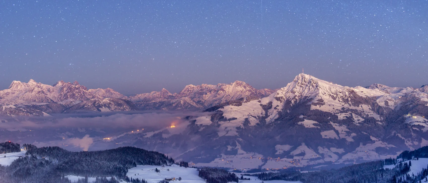 Schneebedeckte Berge mit Sternenhimmel und Lichtern im Tal