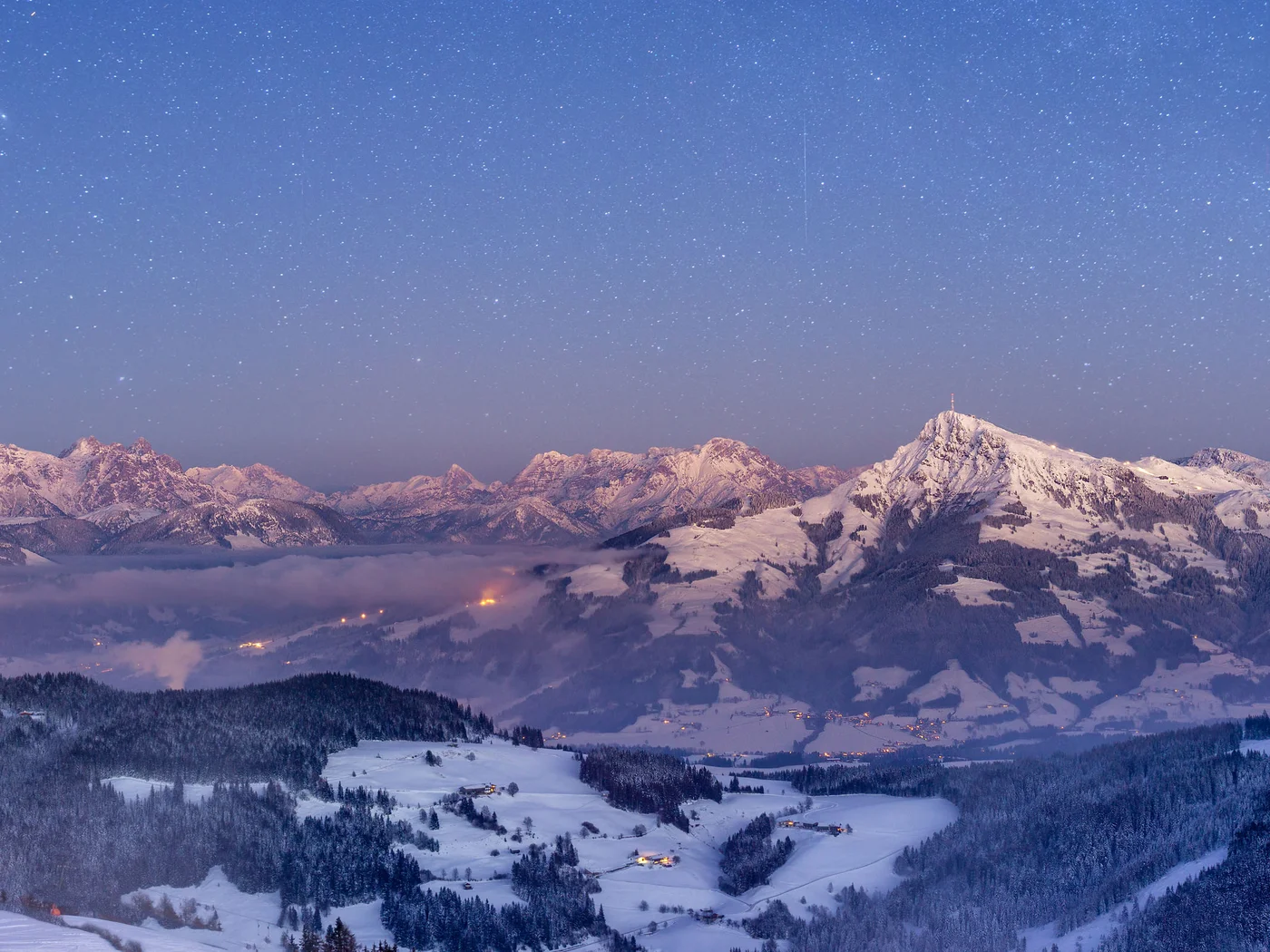 Schneebedeckte Berge mit Sternenhimmel und Lichtern im Tal