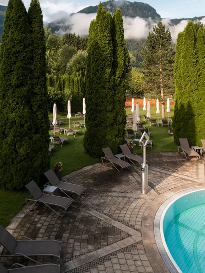 Area piscina con lettini e alberi verdi davanti a montagne sotto un cielo nuvoloso